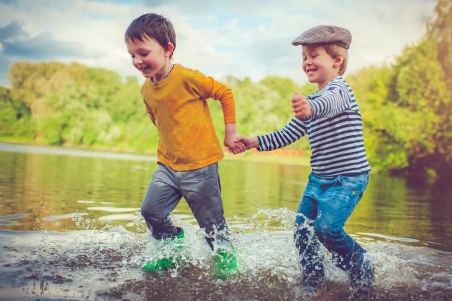 Two young boys are holding hands and running through a puddle of water.