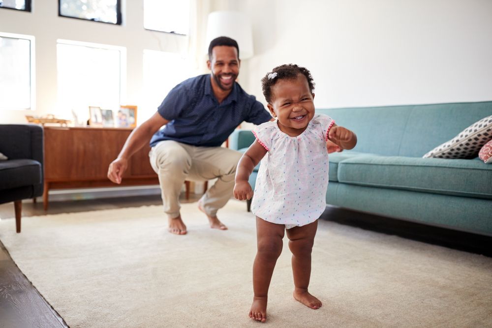 A man is teaching a baby girl how to walk in a living room.