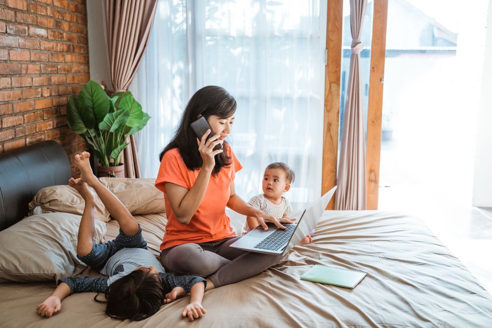 A woman is sitting on a bed using a laptop and talking on a cell phone.