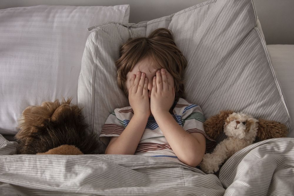 A little boy is laying in bed with a stuffed animal and covering his face with his hands.