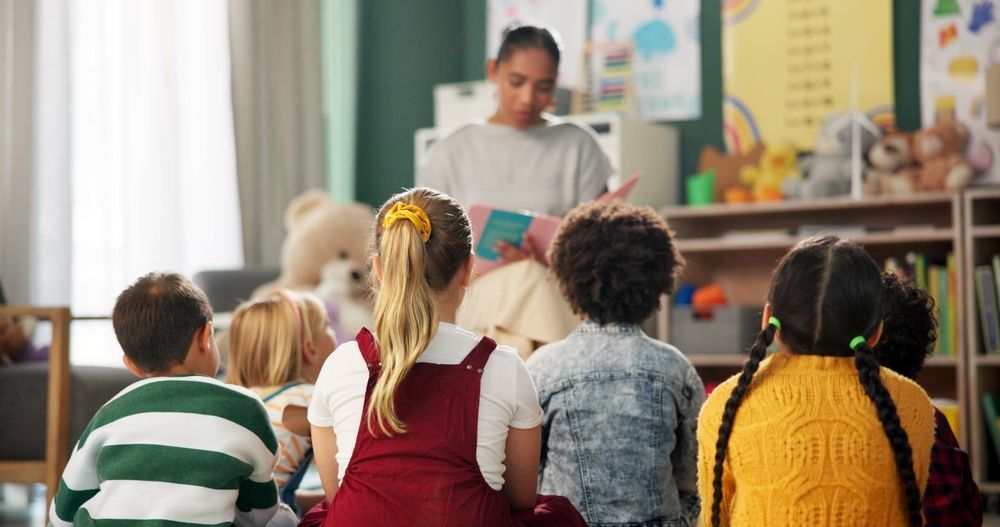 A group of children are sitting on the floor listening to a teacher read a book.