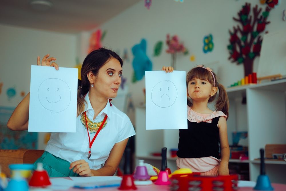 Woman and child holding drawings of smiley and frowny faces, in a colorful classroom.