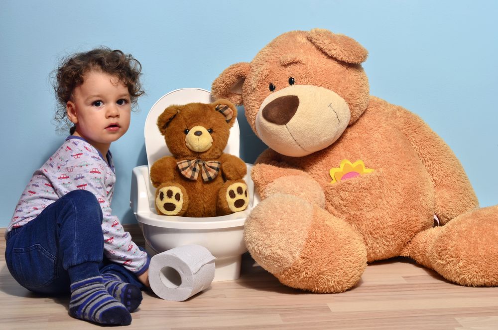 A little girl is sitting on a toilet next to a teddy bear.