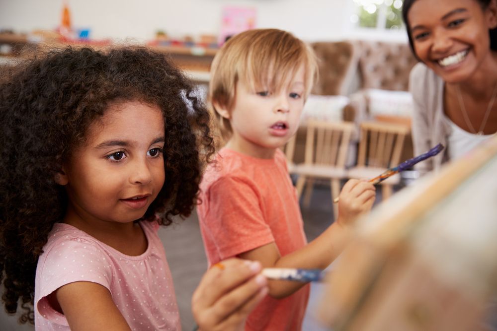 Montessori teacher assessing young students fine motor skills by watching them paint on an easel 