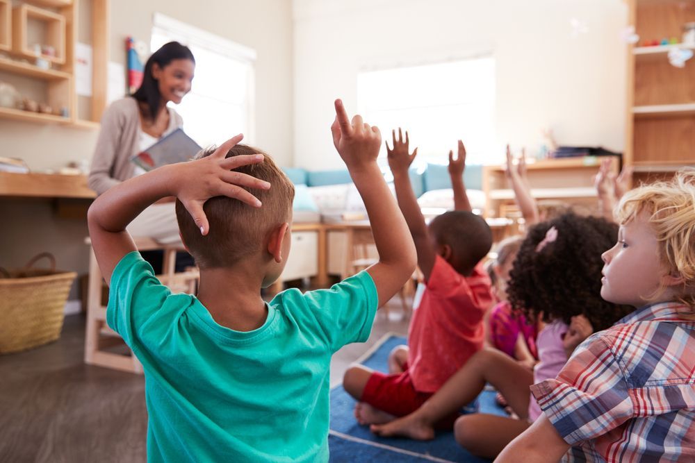 A group of children are sitting on the floor with their hands up in a classroom.