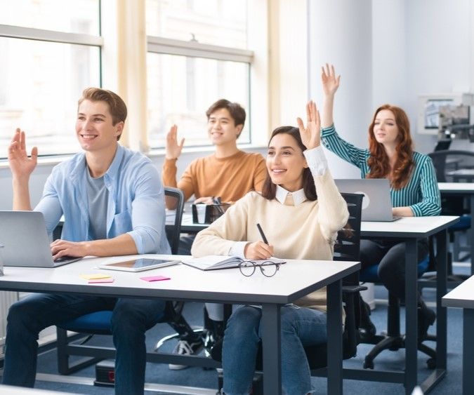 The same young woman sits in her class with her classmates, they are raising their hands to answer a question