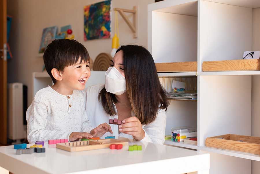 A woman wearing a mask is playing with a child at a table.