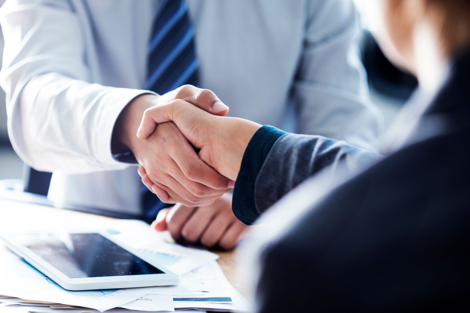 Two people in business attire shaking hands over a table with papers and a tablet.