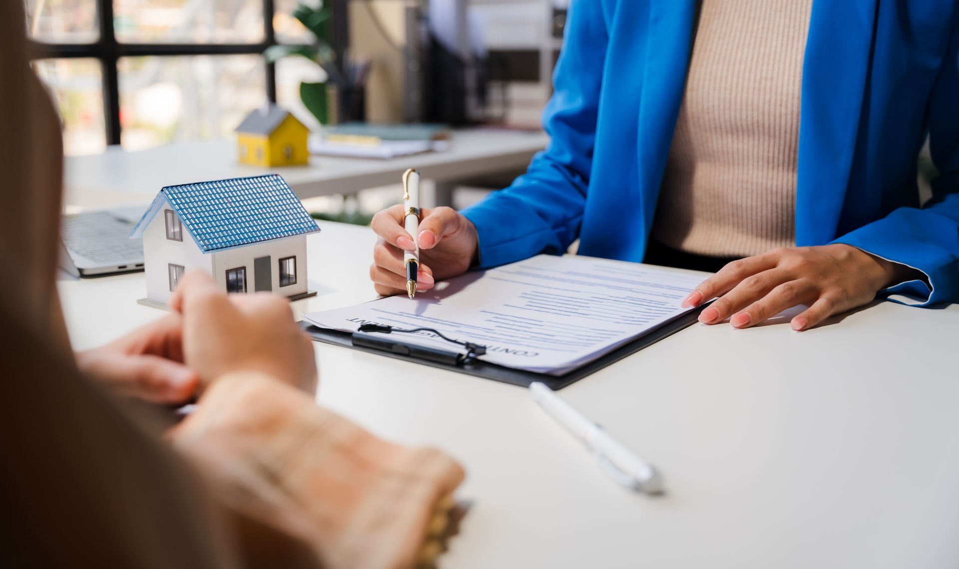 Person signing document with pen, miniature house and other person at table.