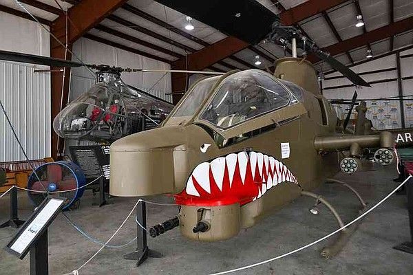 A tan AH-1 Cobra attack helicopter with shark teeth nose art sits inside a museum hangar next to another helicopter.