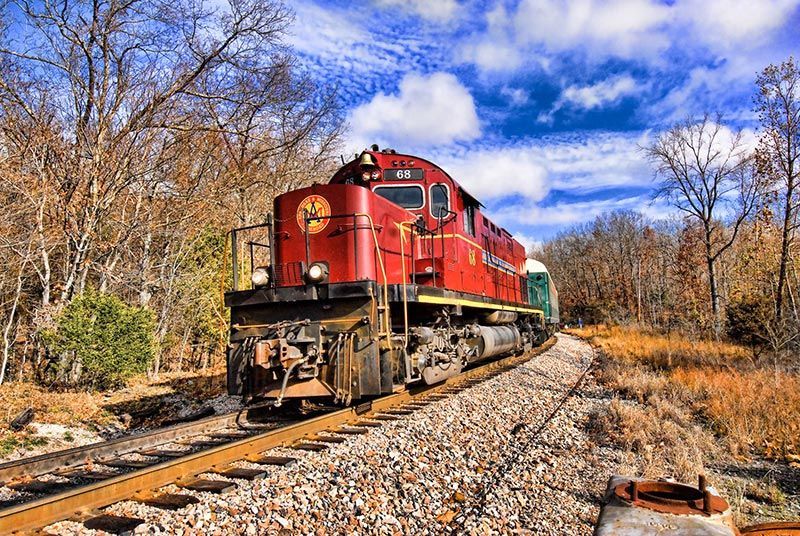 A red locomotive travels on train tracks through a wooded, autumnal landscape under a bright blue sky.