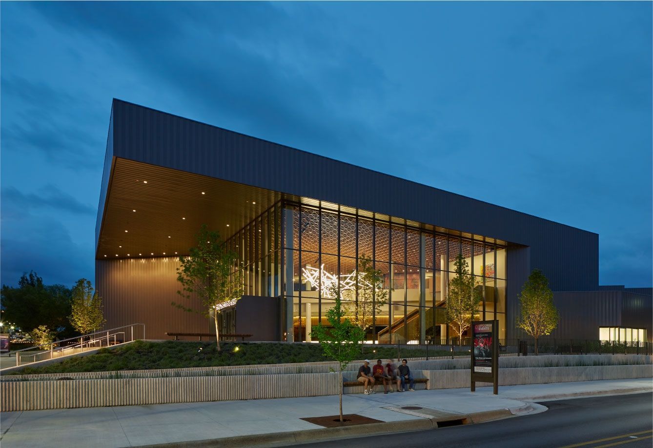 Modern building with dark metal walls, large glass facade, and warm interior lighting, set against a blue twilight sky.