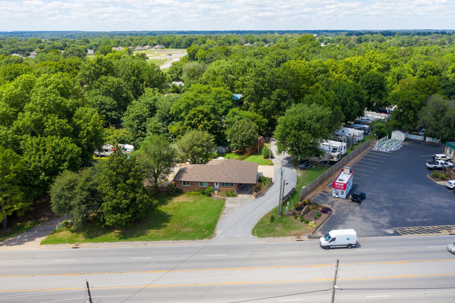 Aerial view of a house and adjacent parking area with RVs next to a road, surrounded by dense trees.