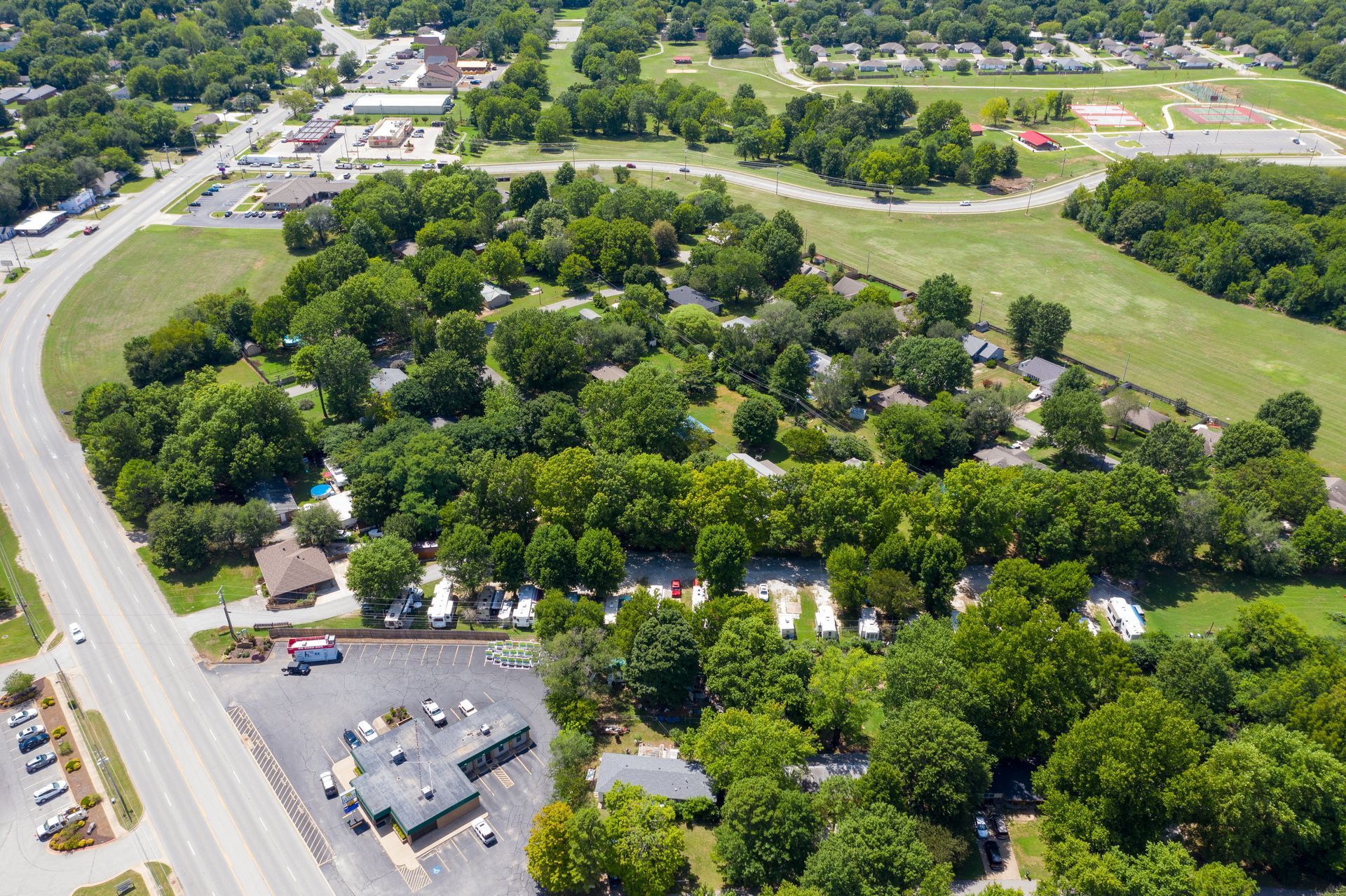An aerial view of a wooded RV park and parking lot located next to a main road in a suburban setting.