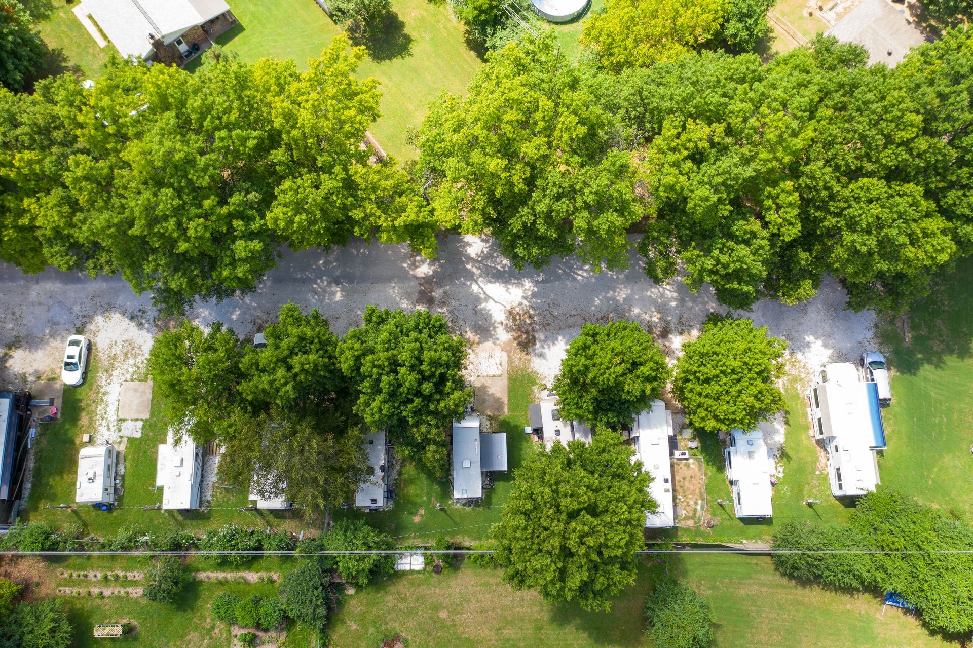 An aerial view of a row of white recreational vehicles parked along a tree-lined gravel road in a grassy area.