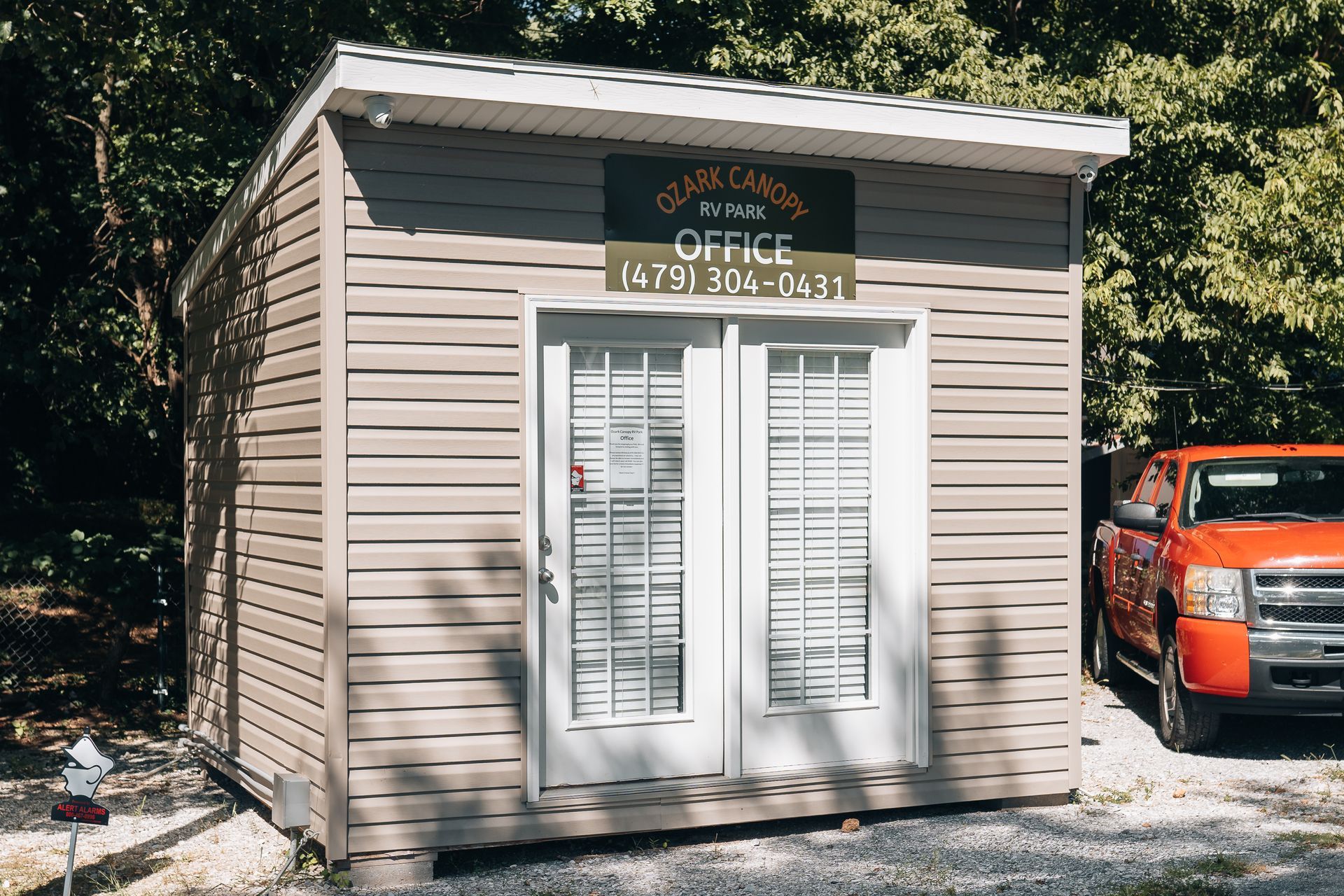 A light brown vinyl-sided shed office with double glass doors, shaded by trees, next to an orange truck.