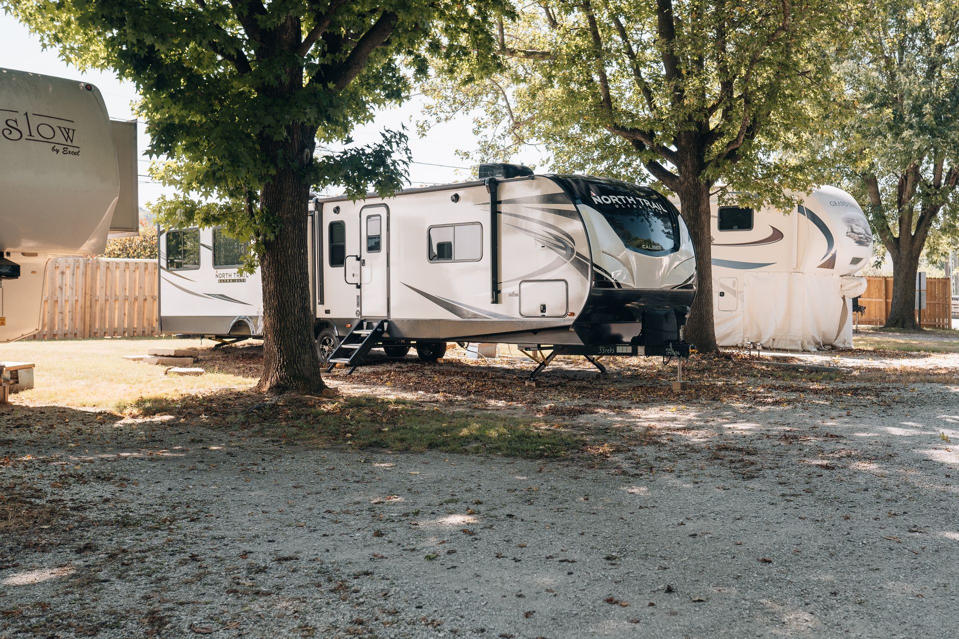 A gravel campground with several recreational vehicles parked under trees on a sunny day.