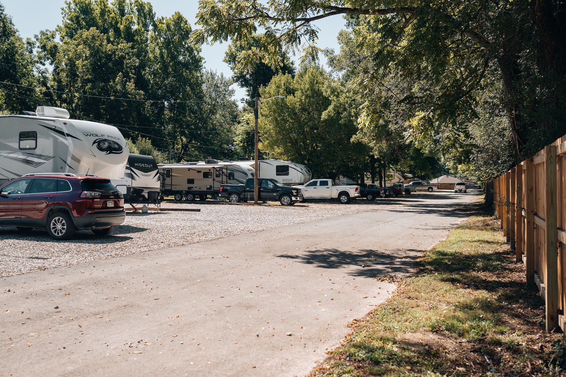 An RV park with several campers and trucks parked on gravel, lined by a wooden fence and trees under a sunny sky.