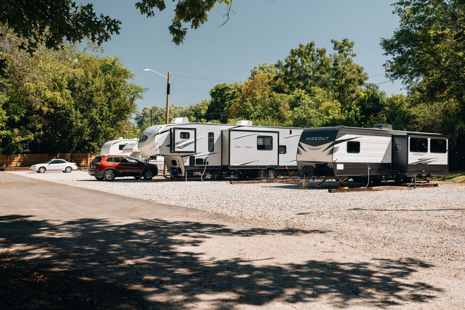 Two recreational vehicles and a car parked on a gravel lot surrounded by trees under a clear blue sky.