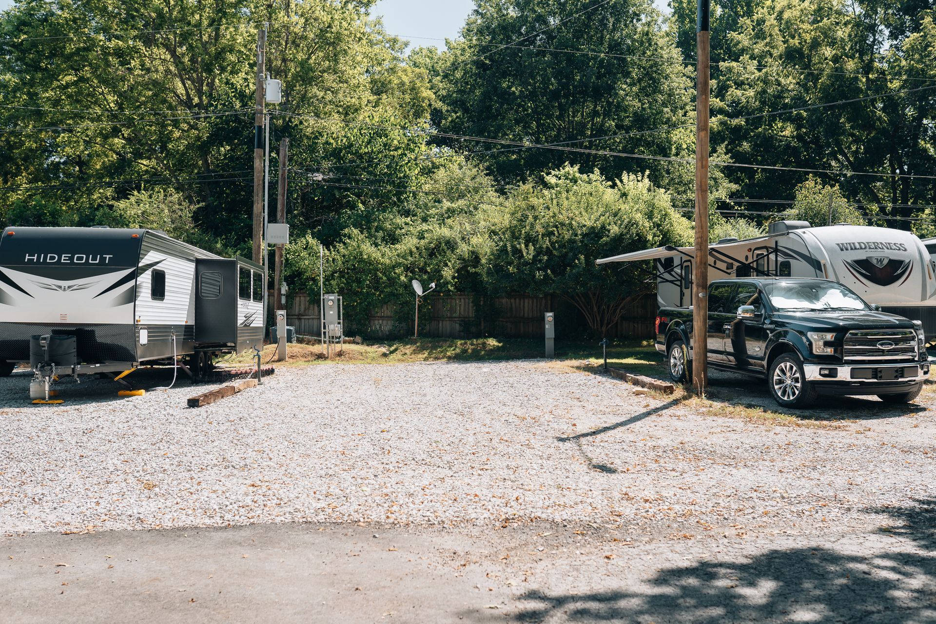 Two travel trailers are parked on a gravel lot in front of a line of dense green trees.