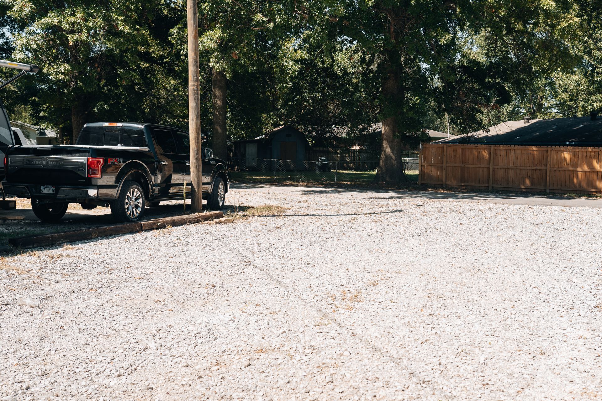 A black pickup truck parked on a gravel lot next to a utility pole, with trees and a wooden fence in the background.