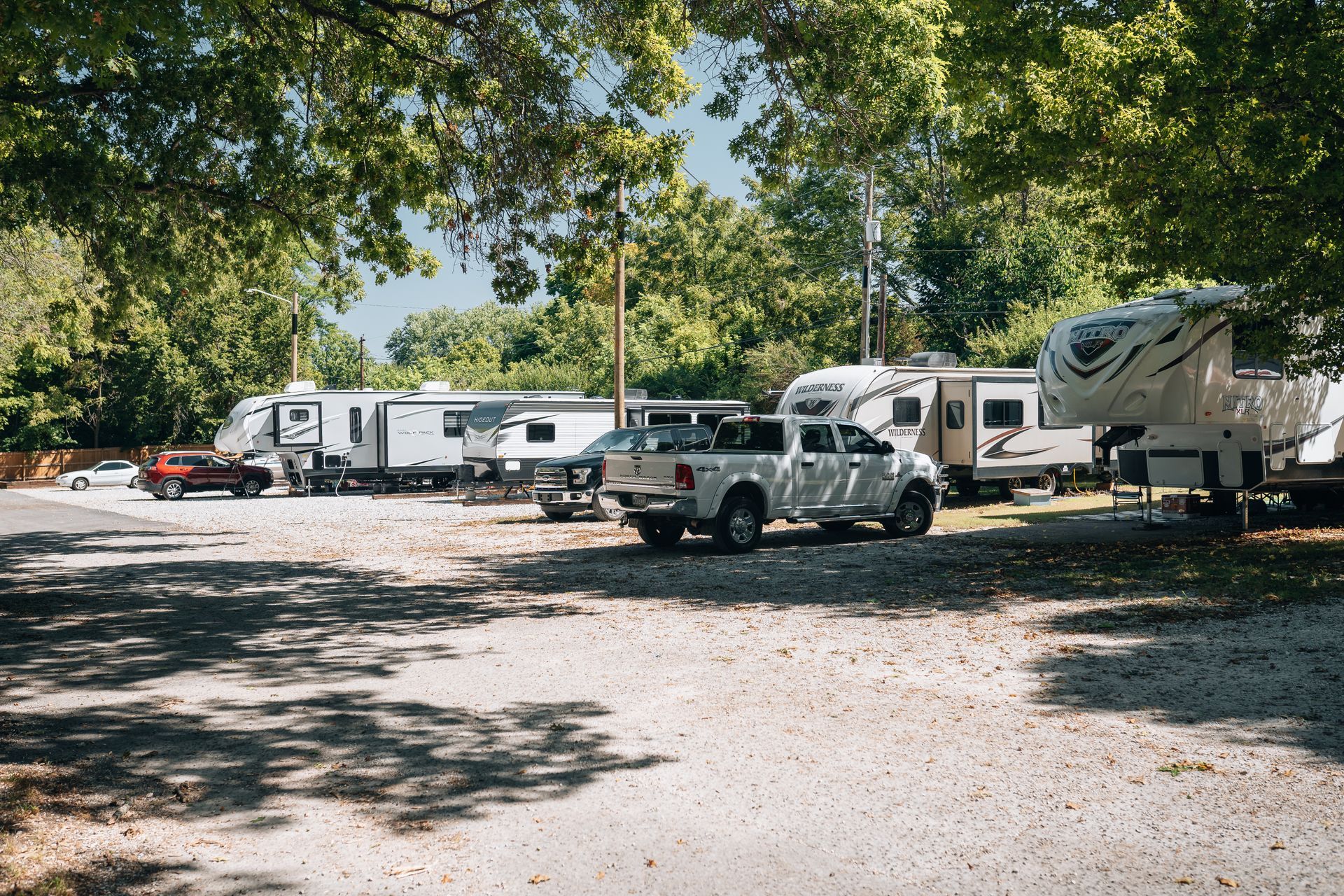 Recreational vehicles and a pickup truck parked on a gravel lot under large, leafy trees on a sunny day.