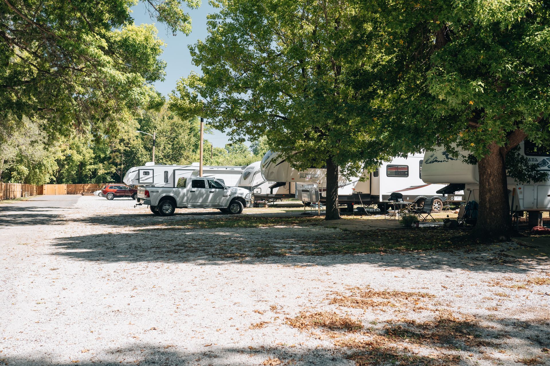 A white pickup truck parked in a gravel campground shaded by large, leafy green trees.