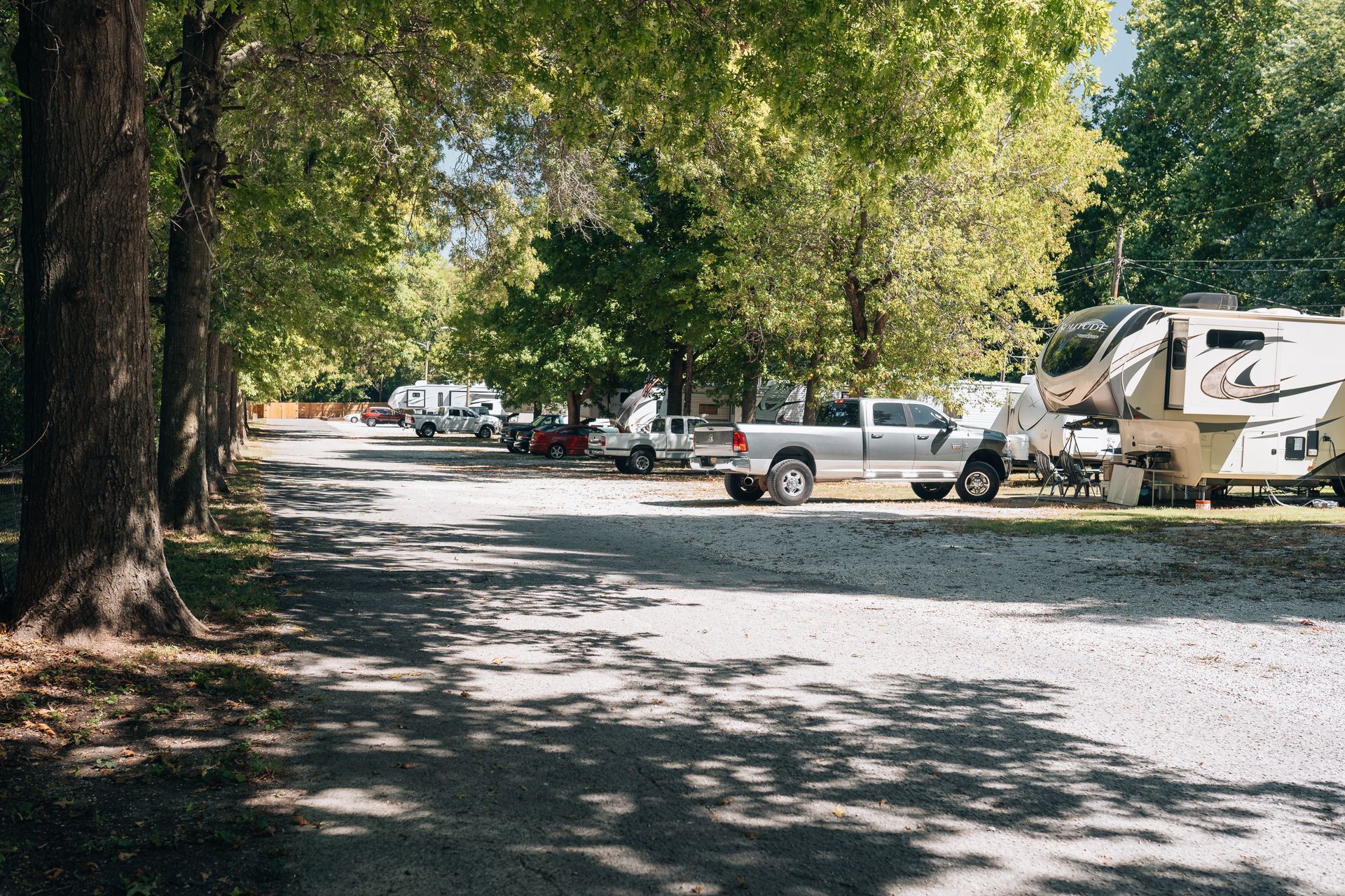 A gravel campground with parked trucks and recreational vehicles under a canopy of green trees on a sunny day.