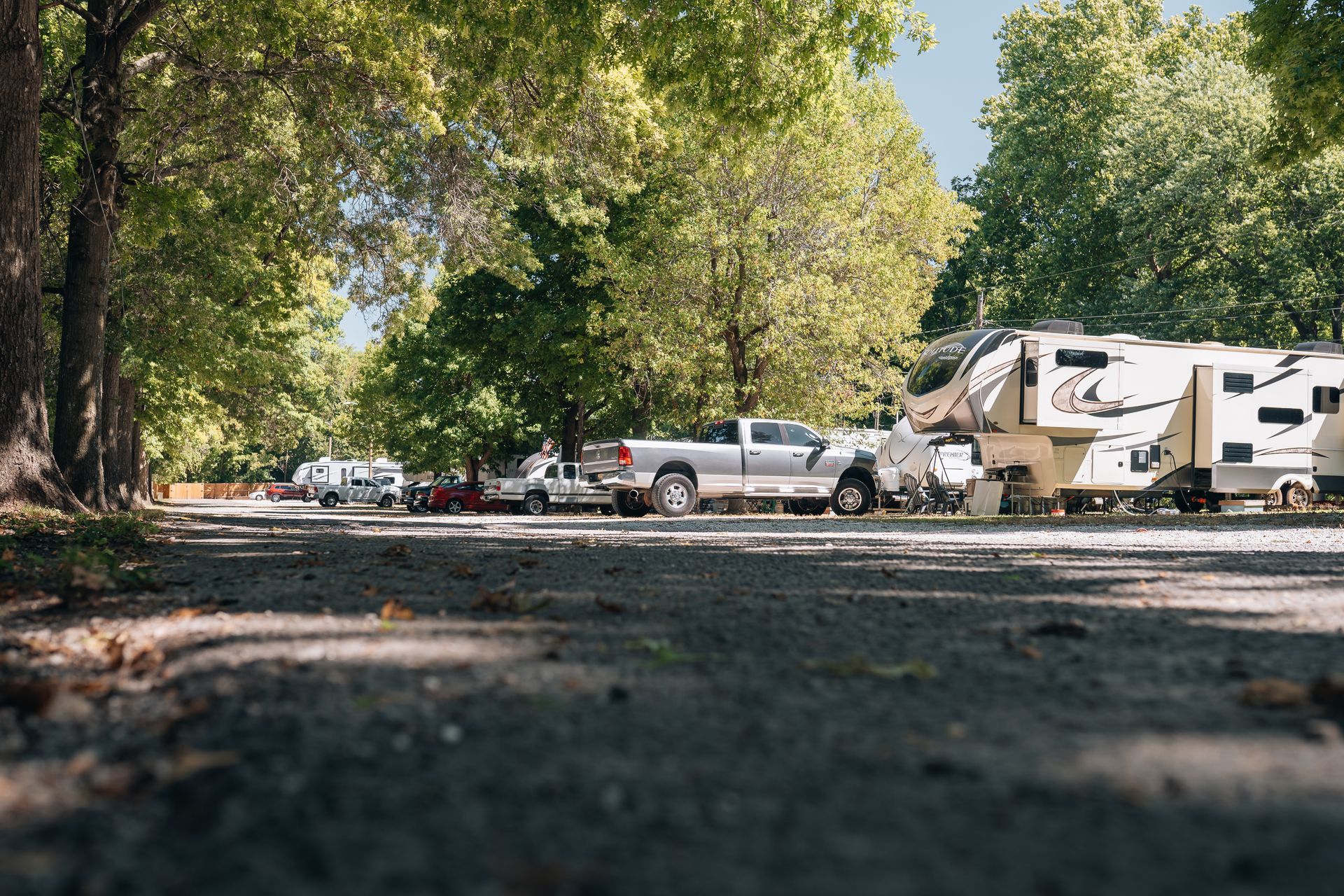 A gravel campground road lined with trees, featuring parked pickup trucks and a large camper trailer in the sunlight.