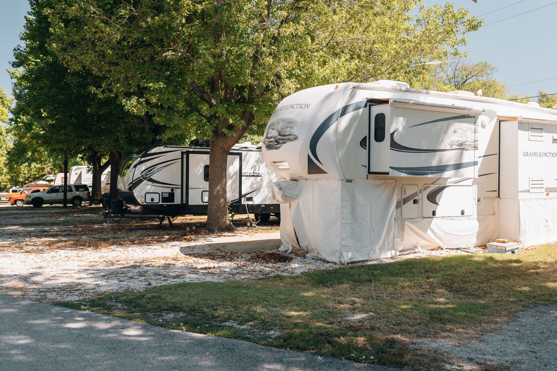 A row of parked recreational vehicles in a shaded, gravel-covered campground during the day.
