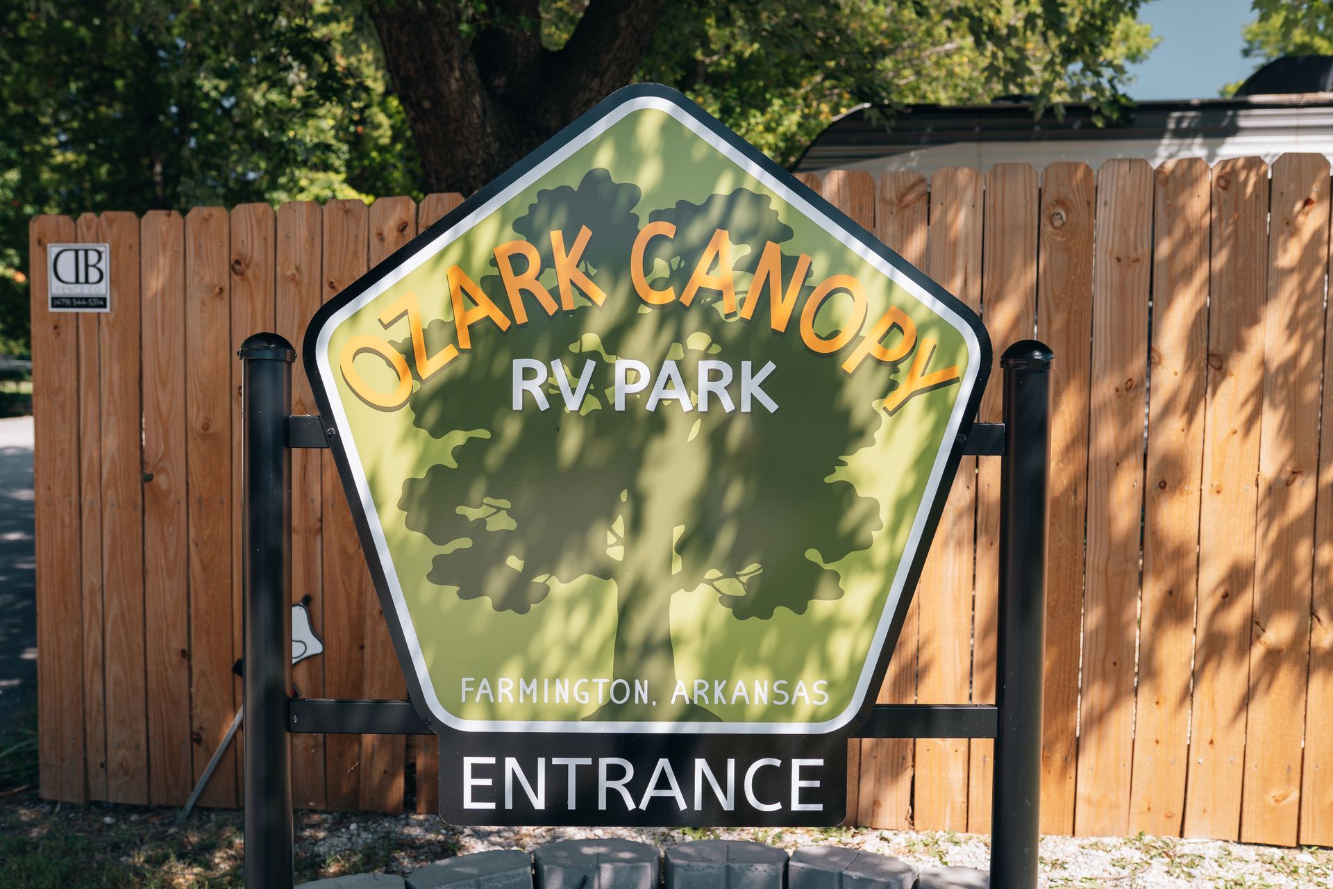 A pentagonal sign for Ozark Canopy RV Park in Farmington, Arkansas, featuring a tree graphic in front of a wood fence.