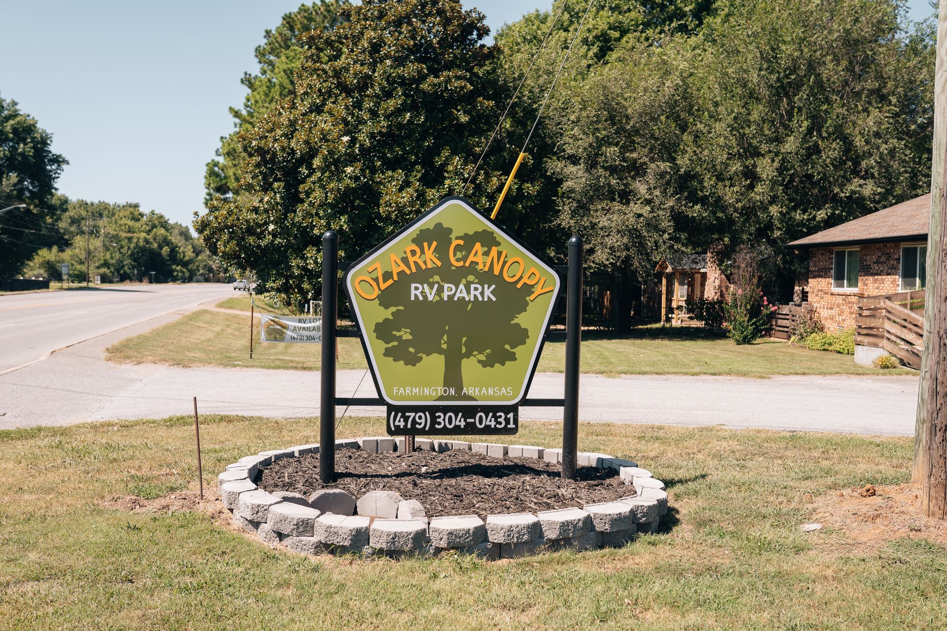 A sign for Ozark Canopy RV Park with a tree graphic, phone number, and decorative stone border in a grassy park setting.