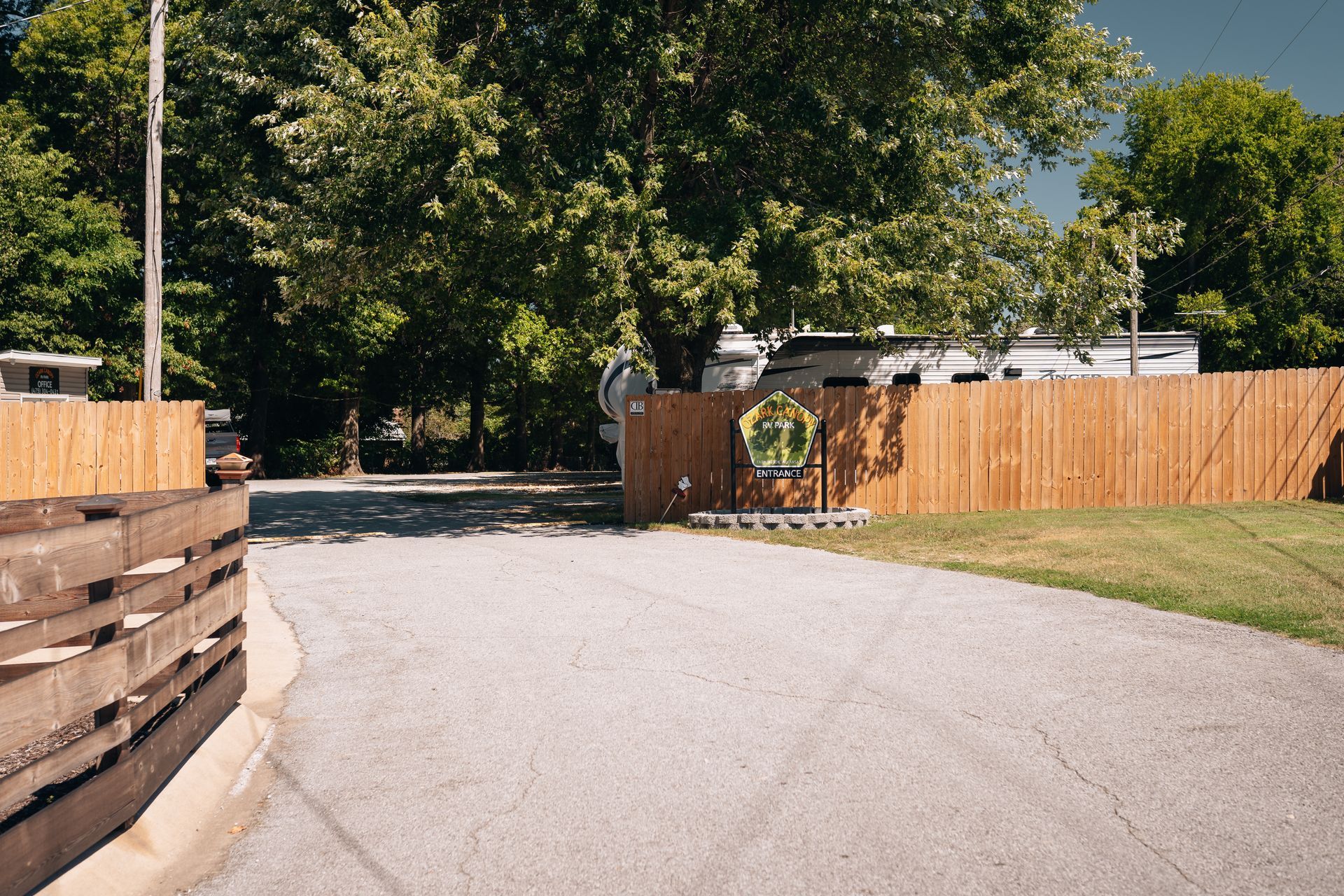 A paved road leads toward a wooden fence and gate, with large trees in the background and an RV partially visible.