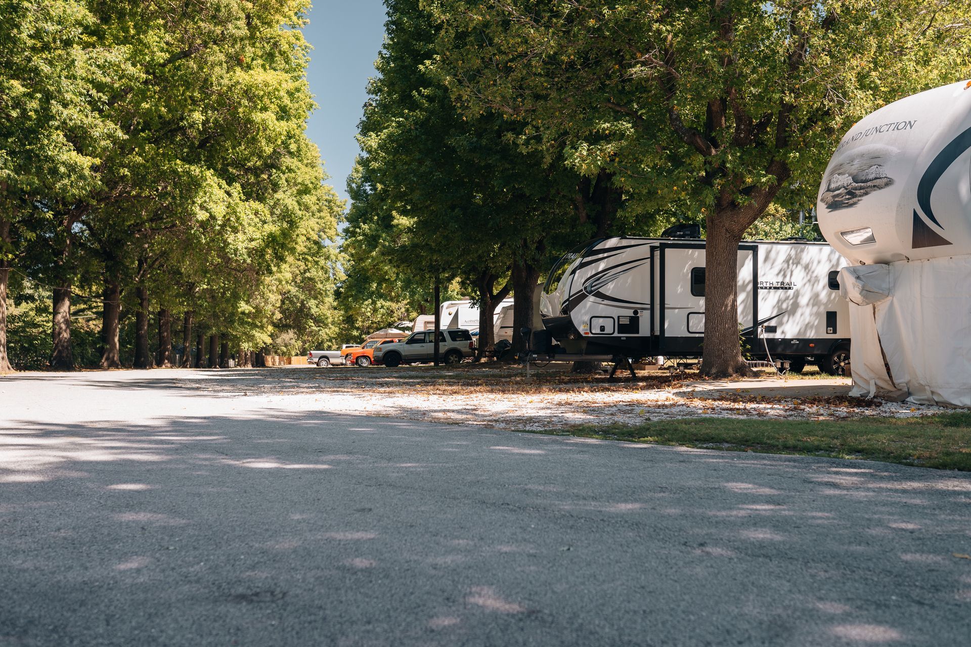 A gravel driveway leads through a shaded campground with parked recreational vehicles under lush green trees.