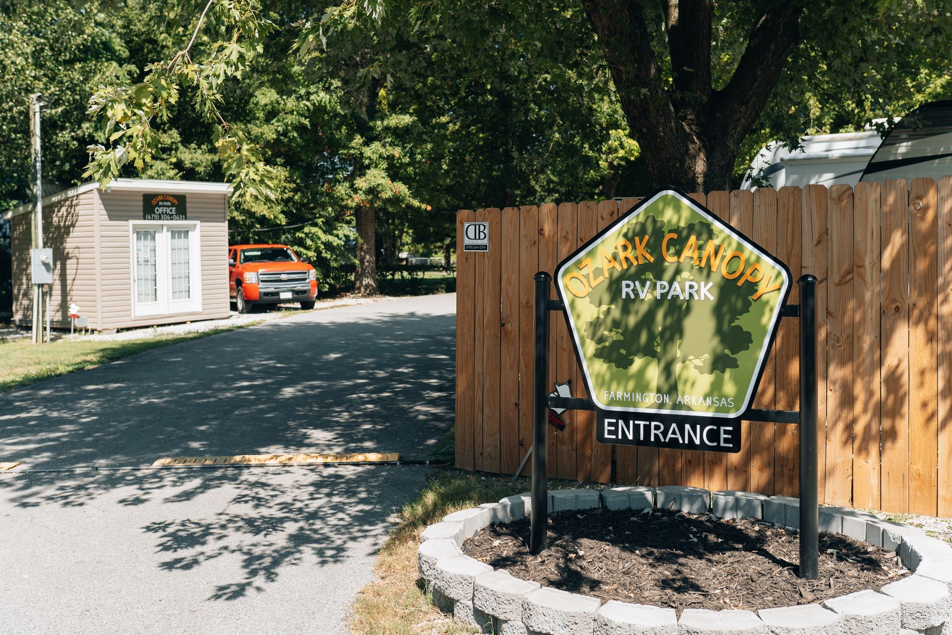 A park entrance sign on a metal stand next to a wooden fence and a small tan building under trees.