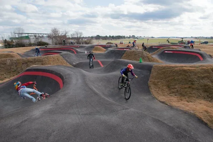 Cyclists ride bikes on an asphalt pump track with rolling hills, curves, and red accents against a cloudy sky.