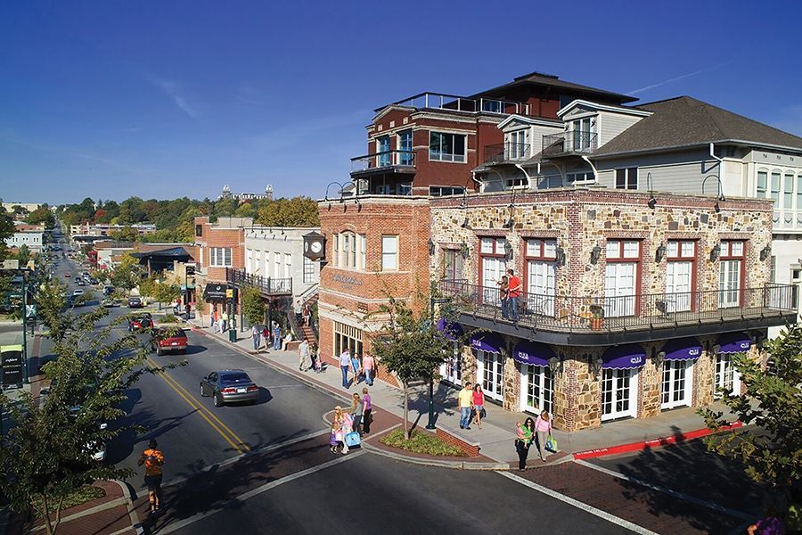 A sunlit city street with pedestrians crossing near a three-story brick and stone building with a balcony and awning.