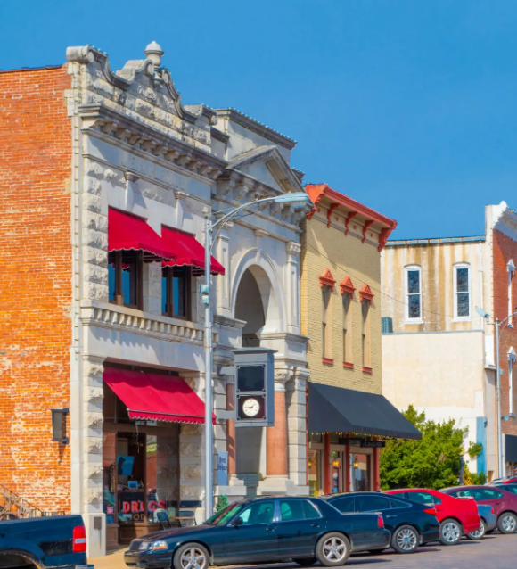A sunny street scene in a historic town with a brick building featuring red awnings, a clock, and parked cars.
