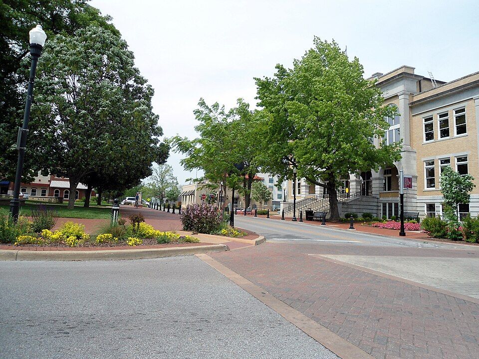 A sunny town square with a brick pathway, mature green trees, and a beige, multi-story government building.