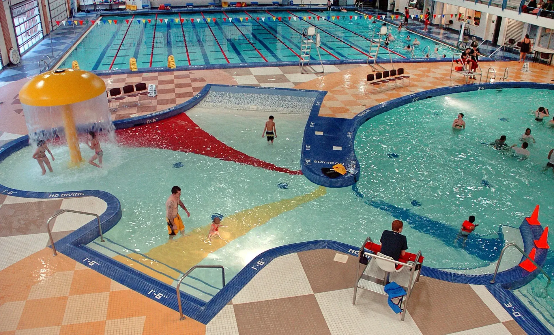 Indoor aquatic center featuring a shallow children's pool with a mushroom fountain and a larger lap pool in the background.