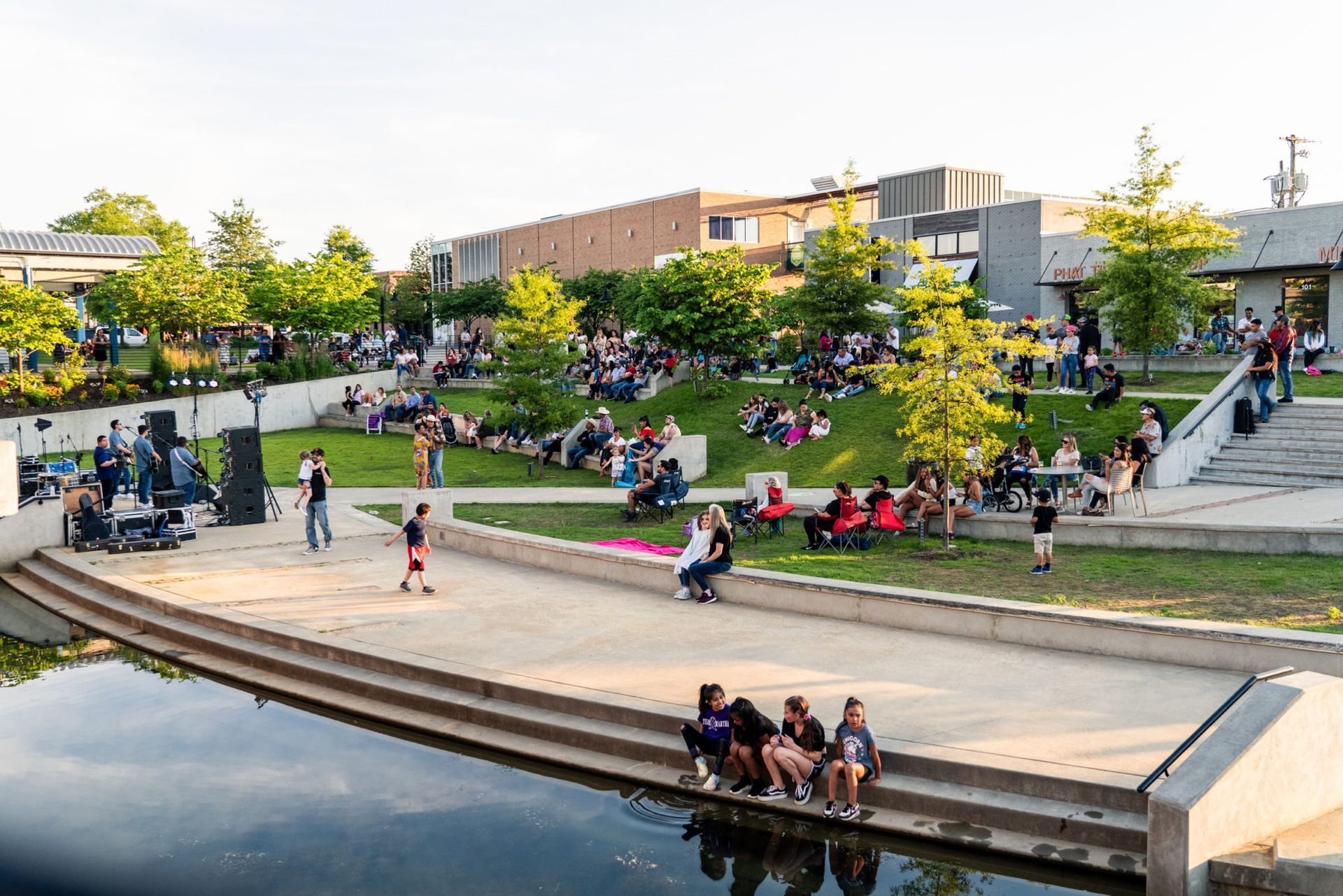 People sit on terraced concrete steps by a canal in a park during a sunny outdoor event with trees and distant buildings.