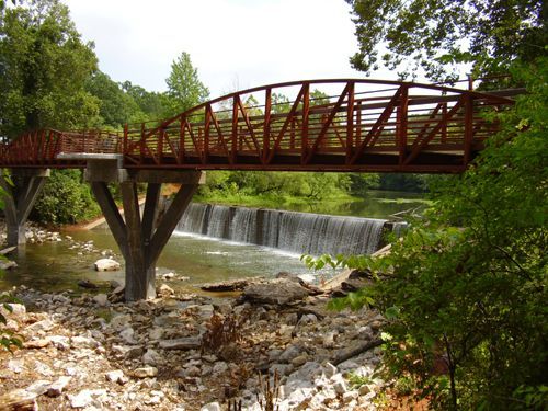A rusted metal pedestrian bridge crosses a stream above a small waterfall, surrounded by lush trees and rocky banks.