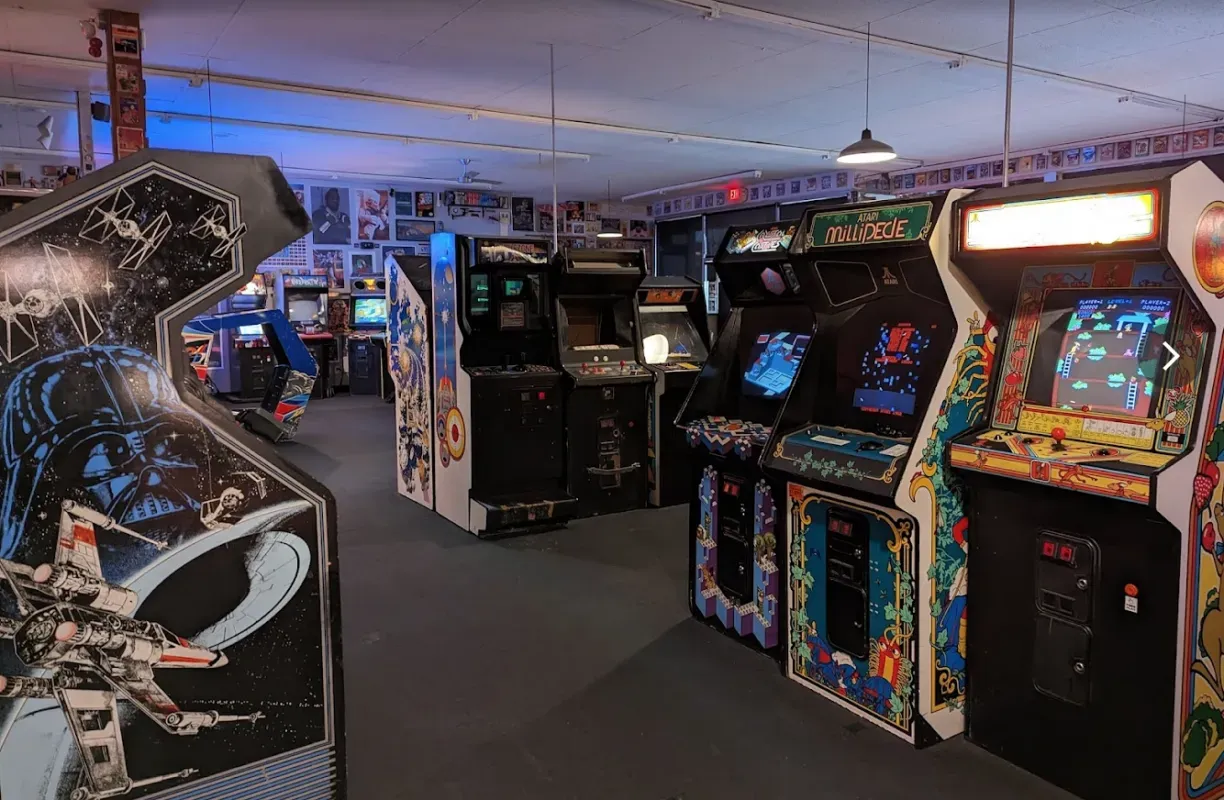 A dimly lit arcade room with rows of colorful classic video game cabinets arranged in a line.