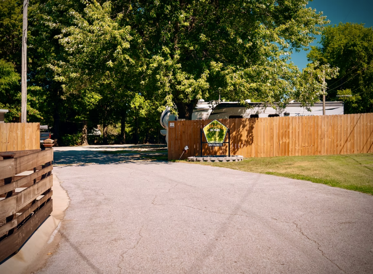 A paved driveway leads to a wooden fence with a small, shield-shaped park sign, surrounded by lush green trees.