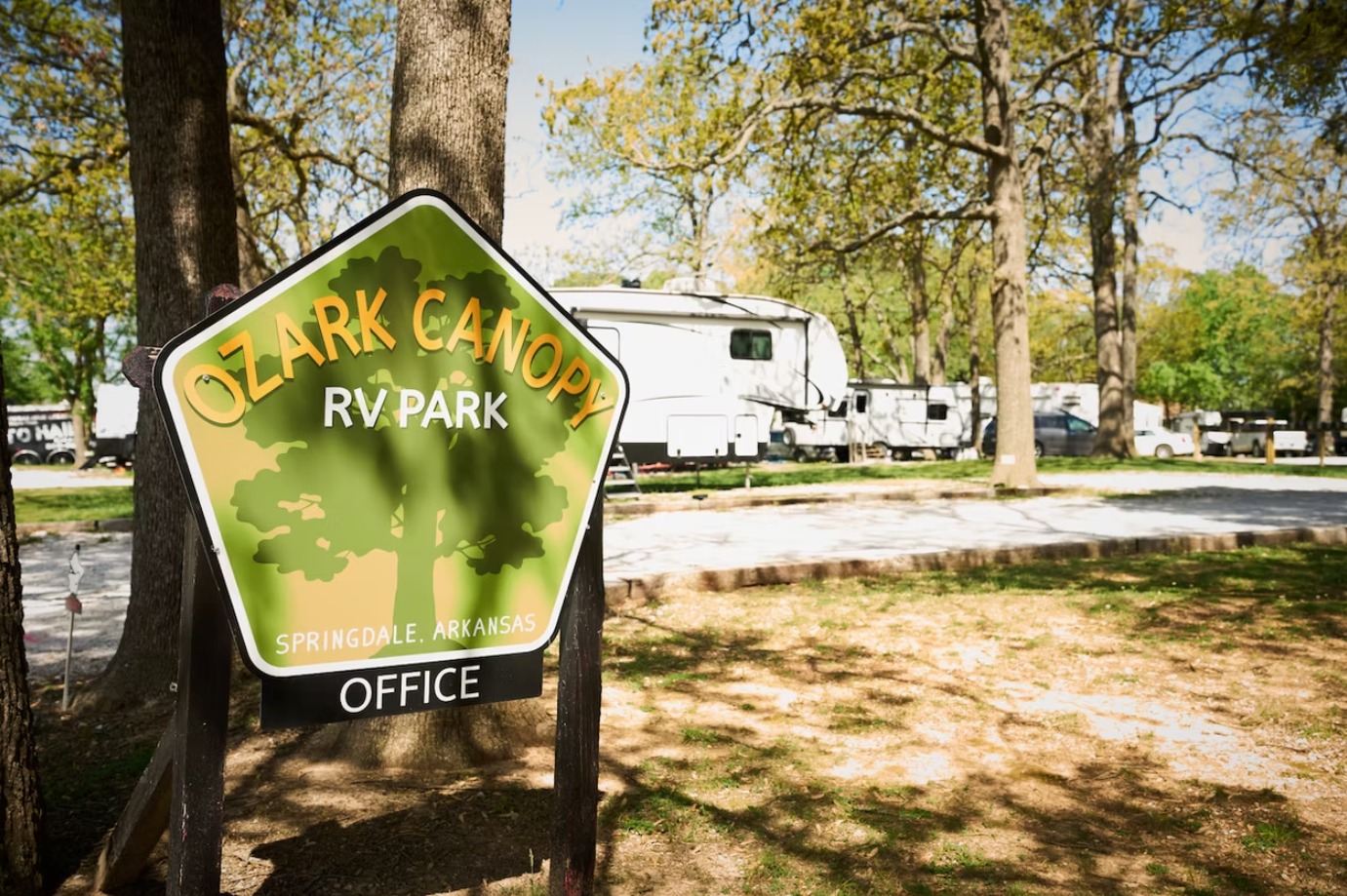 A pentagonal Ozark Canopy RV Park sign stands in a wooded campground with white RVs parked in the background.
