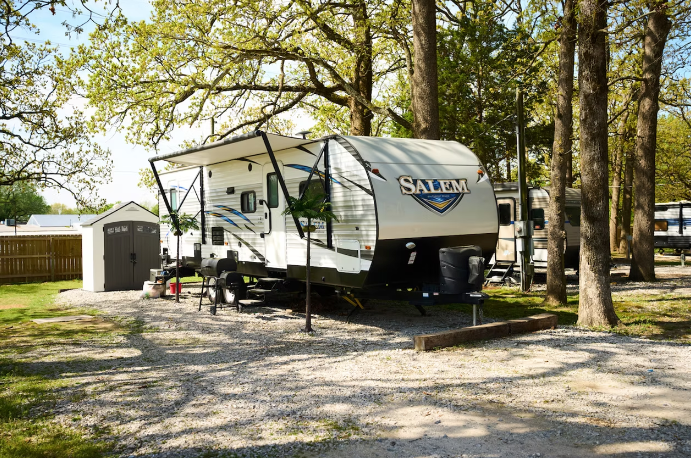 A white Salem travel trailer parked in a gravel lot under large trees next to a small, dark storage shed.