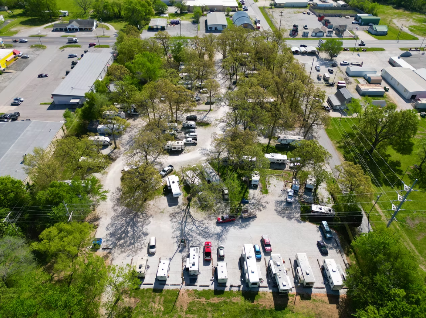An aerial view of an RV park with gravel paths, mature trees, and numerous campers and vehicles parked in designated spots.