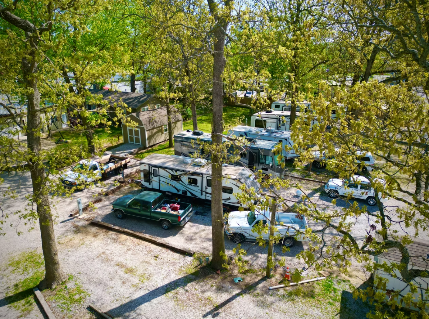 An aerial view of a wooded RV park with several campers, pickup trucks, and a small building among leafy trees.