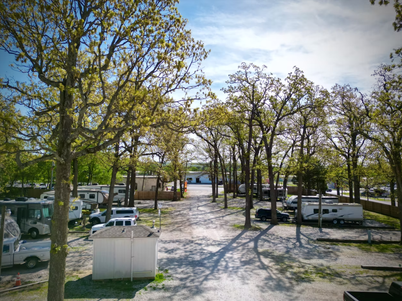A gravel path leads through a wooded RV park with parked vehicles and campers under bright, sunlit trees.