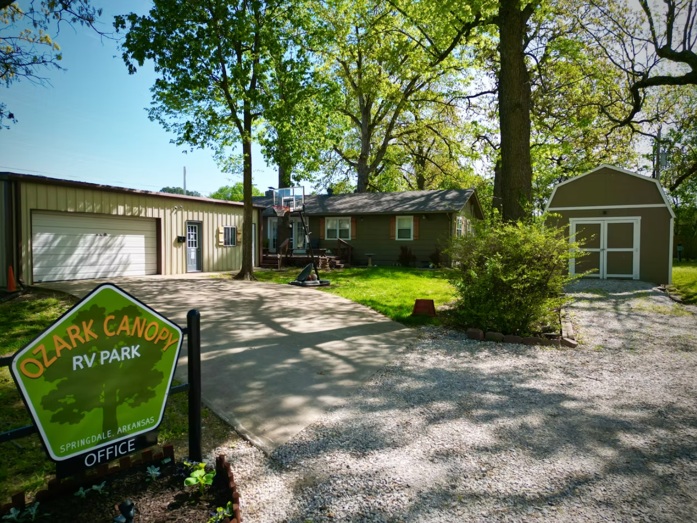 A green sign for Ozark Canopy RV Park stands before a driveway, a detached garage, a house, and a shed on a sunny day.